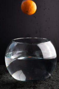 scenario for water splash photo. glass aquarium with water, black backdrops. orange fruit still in the air going into aquarium direction