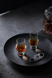 2 glasses of whisky on a black tray with a cigar cutter. dark and sophisticated ambient with back side light