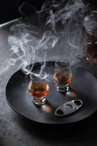 2 glasses of whisky on a black tray with a cigar cutter. dark and sophisticated ambient, smoke and back side light