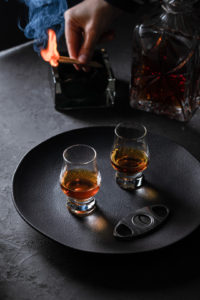 2 glasses of whisky on a black tray in a dark sophisticated ambient. lit matches with smoke